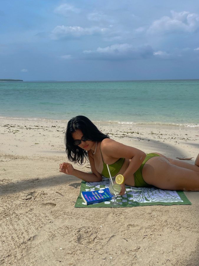 Girl in a bikini laying on a blanket on a sandy beach with a drink and a book